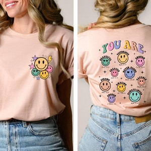 K&ouml;nnte beinhalten: Ein rosa T-Shirt mit einem Grafikdruck von Smiley-Gesichtern und Sternen auf der Vorderseite und dem Text "YOU ARE" mit einem Grafikdruck von Smiley-Gesichtern und Bibelversen auf der R&uuml;ckseite.