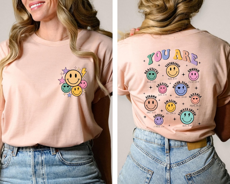 K&ouml;nnte beinhalten: Ein rosa T-Shirt mit einem Grafikdruck von Smiley-Gesichtern und Sternen auf der Vorderseite und dem Text "YOU ARE" mit einem Grafikdruck von Smiley-Gesichtern und Bibelversen auf der R&uuml;ckseite.