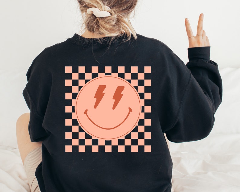 May include: Black sweatshirt with a pink smiley face with lightning bolts for eyes on a checkered background.