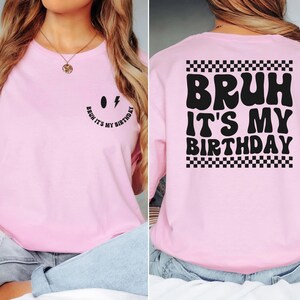 May include: Pink t-shirt with a black smiley face on the front and the text "Bruh it's my birthday" in a retro font with a checkered border on the back.