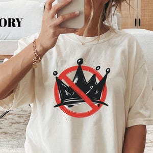 Pode incluir: Camiseta cor marfim com um desenho de coroa preta riscada com um c&iacute;rculo vermelho. A camiseta gr&aacute;fica tem um ajuste folgado e mangas curtas. A palavra "IVORY" &eacute; impressa no topo.