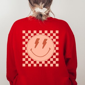 May include: Red sweatshirt featuring a checkered pattern and a peach-coloured smiley face with lightning bolts for eyes.
