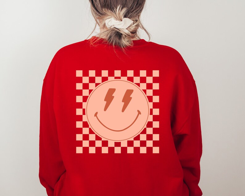 May include: Red sweatshirt featuring a checkered pattern and a peach-coloured smiley face with lightning bolts for eyes.