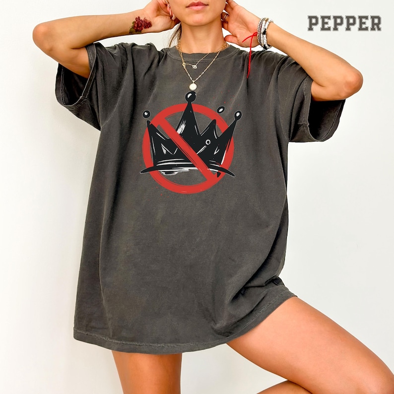 Pode incluir: Camiseta cinza escuro com um desenho de coroa preta riscada com um c&iacute;rculo e uma linha vermelhos. A palavra "PEPPER" est&aacute; impressa em branco no canto superior direito. A camiseta tem mangas curtas e um corte folgado.