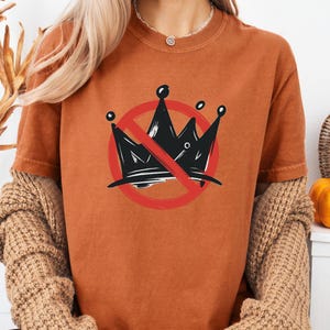 Pode incluir: Camiseta cor de ferrugem com um desenho de coroa preta dentro de um c&iacute;rculo vermelho com uma linha a atravess&aacute;-lo. A palavra "YAM" est&aacute; impressa no canto superior esquerdo. A camisa tem mangas curtas e gola redonda.