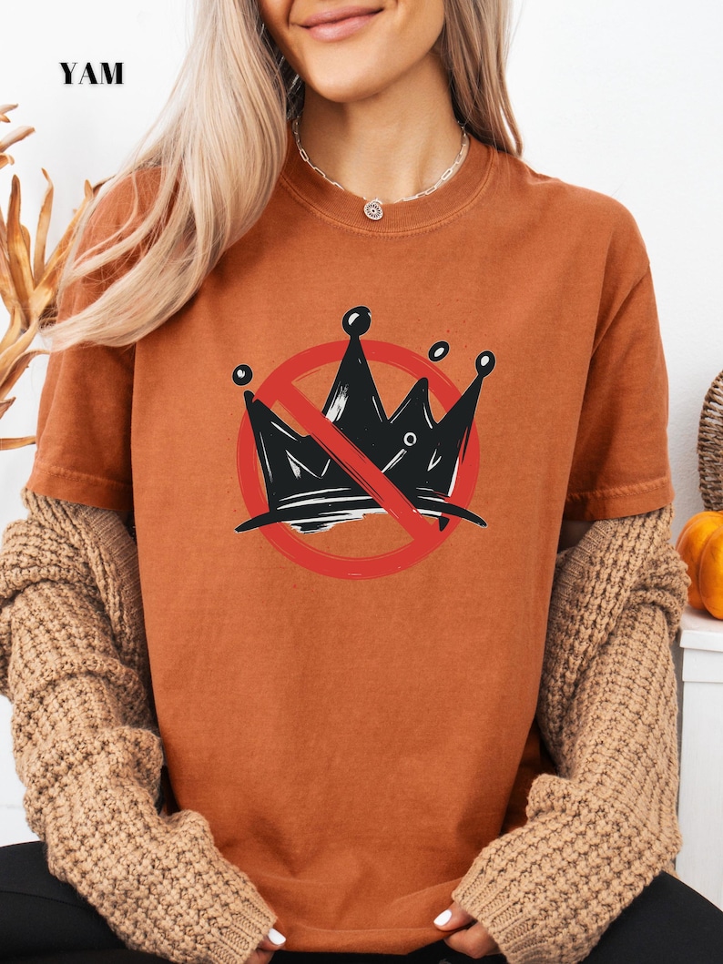 Pode incluir: Camiseta cor de ferrugem com um desenho de coroa preta dentro de um c&iacute;rculo vermelho com uma linha a atravess&aacute;-lo. A palavra "YAM" est&aacute; impressa no canto superior esquerdo. A camisa tem mangas curtas e gola redonda.