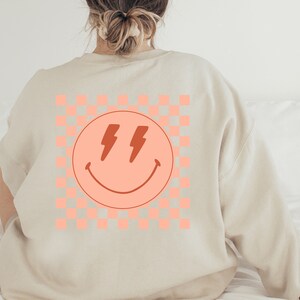 May include: A beige sweatshirt with a pink and white chequered pattern and a pink smiley face with red lightning bolts for eyes.