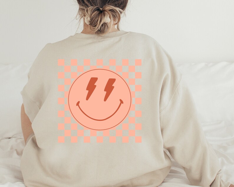 May include: A beige sweatshirt with a pink and white chequered pattern and a pink smiley face with red lightning bolts for eyes.