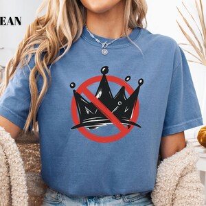 Pode incluir: Camiseta azul jeans com um gr&aacute;fico de uma coroa preta riscada por um c&iacute;rculo vermelho e uma linha diagonal. O texto "BLUE JEAN" est&aacute; no canto superior esquerdo.