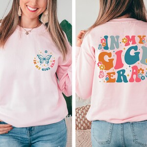 May include: Pink sweatshirt with a butterfly and the text "In My Gigi Era" on the front and the same text in larger, colorful letters on the back.