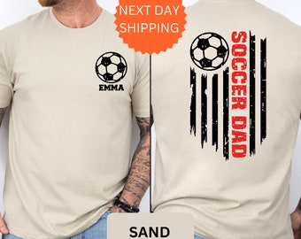 May include: Sand-colored t-shirt with a soccer ball and the name "Emma" on the front. The back of the shirt features a distressed American flag with the words "Soccer Dad" in red.