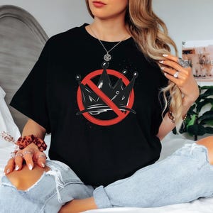 Pode incluir: T-shirt preto com um gr&aacute;fico de uma coroa com um c&iacute;rculo vermelho e uma linha vermelha a atravess&aacute;-la. O gr&aacute;fico est&aacute; centrado na frente da t-shirt. A t-shirt &eacute; usada por uma pessoa sentada numa cama.
