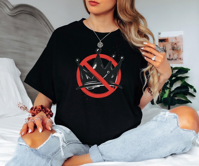 Pode incluir: T-shirt preto com um gr&aacute;fico de uma coroa com um c&iacute;rculo vermelho e uma linha vermelha a atravess&aacute;-la. O gr&aacute;fico est&aacute; centrado na frente da t-shirt. A t-shirt &eacute; usada por uma pessoa sentada numa cama.