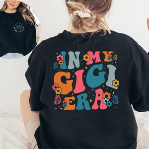 May include: Black sweatshirt with a colorful graphic design that says "In My Gigi Era" with flowers and butterflies.