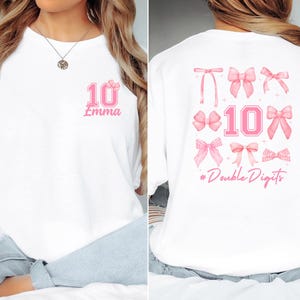 May include: White t-shirt with pink graphics. The front features the number "10" with a bow and the name "Emma". The back displays various pink bows, the number "10", and the phrase "#Double Digits".