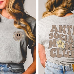 May include: A gray t-shirt with a brown smiley face with flowers around it on the front and the text "Anti Social Moms Club" in brown with a yellow flower on the back.