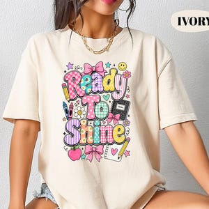 May include: An ivory short-sleeve t-shirt featuring a colorful graphic print with the text Ready To Shine. The design includes school-themed icons like a notebook, pencils, crayons, an apple, and bows in shades of pink, blue, yellow, and purple.