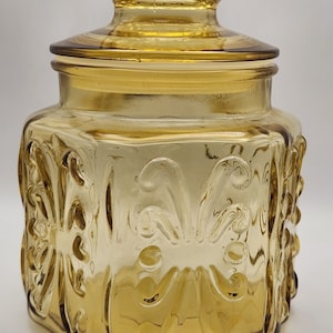 Puede incluir: Un tarro de vidrio ámbar vintage con tapa. El tarro tiene forma hexagonal y está decorado con un patrón floral en relieve.