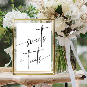 May include: A gold-framed sign with the text "sweets + treats" written in black calligraphy. The sign is sitting on a table with a white bouquet of flowers in a glass vase.
