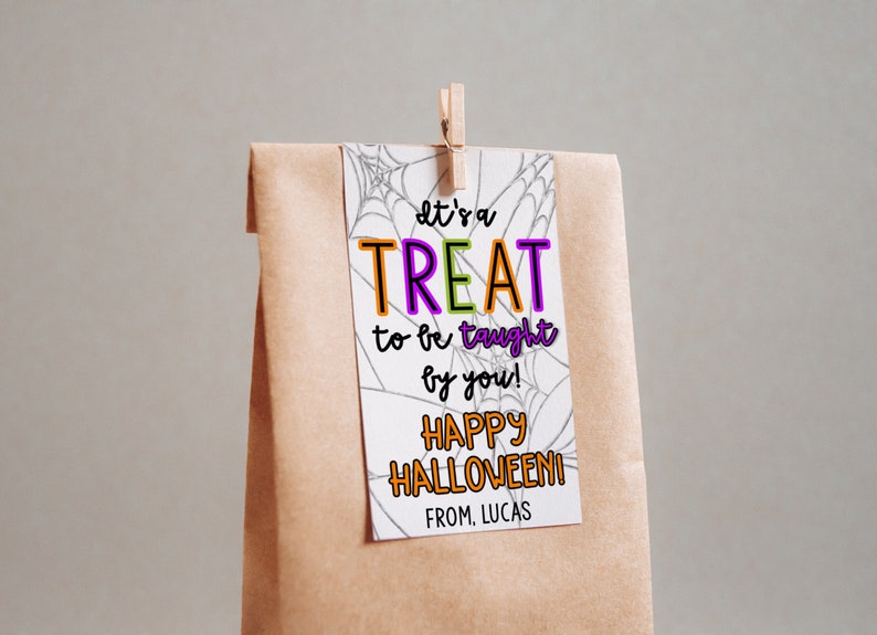 Puede incluir: Una tarjeta blanca con un dise&ntilde;o de telara&ntilde;a negra y el texto "It's a TREAT to be taught by you! HAPPY HALLOWEEN! FROM. LUCAS" en letras de colores. La tarjeta est&aacute; unida a una bolsa de papel marr&oacute;n con una pinza de ropa.