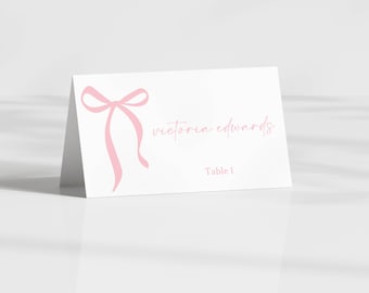May include: A white place card with a pink bow illustration and the name "victoria edwards" in a cursive font. The text "Table 1" is printed below the name. The card is angled, suggesting it is standing.