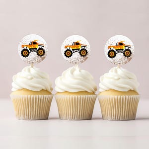 May include: Three cupcakes decorated with orange and black monster truck toppers. The cupcakes are frosted with white frosting and have a white paper liner.