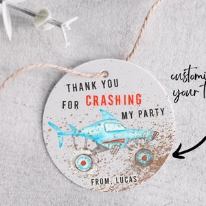 May include: Round thank you tag with a blue and white monster truck with shark fins. The tag has the text "Thank you for crashing my party" and "From, Lucas".