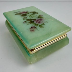 Peut inclure: Une boîte rectangulaire en pierre vert clair avec un couvercle à charnière orné d'un motif floral peint représentant des roses blanches et roses avec des feuilles vertes. La boîte a une finition polie et une bordure en métal doré.