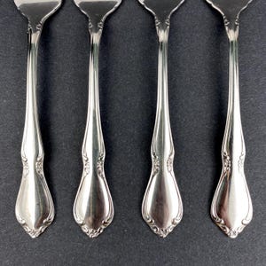 May include: Four silver forks with decorative floral patterns on the handles and tines. The forks are displayed in a row against a dark backdrop, highlighting their polished finish and elegant design. The forks are a classic style.