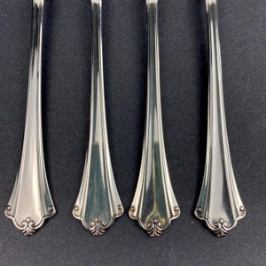 May include: Four silver-coloured forks with a decorative floral design at the base of the tines. The forks have a polished, reflective surface, and the handles taper to a point. The background is a dark, solid colour.