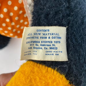 以下が含まれることがあります： タグが付いたヴィンテージのぬいぐるみ。タグには「CONTENTS ALL NEW MATERIAL SYNTHETIC FOAM & COTTON CALIFORNIA STUFFED TOYS 611 So. Anderson St. Los Angeles, Ca. 90023」と書かれています。おもちゃは黒、黄色、オレンジ色の生地です。