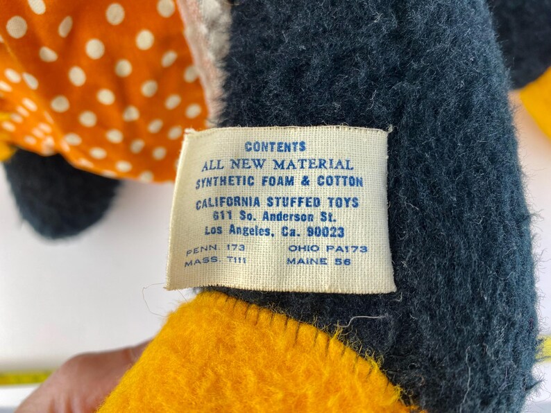 以下が含まれることがあります： タグが付いたヴィンテージのぬいぐるみ。タグには「CONTENTS ALL NEW MATERIAL SYNTHETIC FOAM & COTTON CALIFORNIA STUFFED TOYS 611 So. Anderson St. Los Angeles, Ca. 90023」と書かれています。おもちゃは黒、黄色、オレンジ色の生地です。