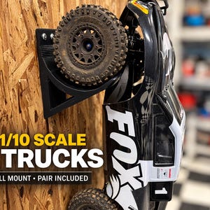 May include: Black wall mount holding a black and white RC truck. The mount also holds a tire. The text on the image says "FITS 1/10 SCALE RC TRUCKS" and "STRONG WALL MOUNT • PAIR INCLUDED". The background is a wooden wall.