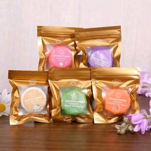 May include: Five gold-colored pouches containing round shower steamers. The labels read Rose, Lavender, Jasmine, Mint & Eucalyptus, and Grapefruit & Ginger. Each pouch is 30g and features a clear window displaying the colored steamer inside.