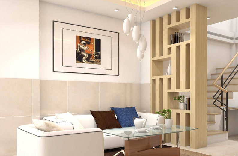 May include: A white sectional sofa with brown and blue throw pillows sits in front of a glass coffee table. A wooden room divider with shelves is to the right of the sofa. A framed picture of an astronaut sitting on a chair is hanging on the wall.
