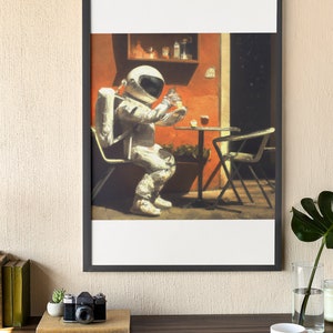 May include: A framed illustration of an astronaut sitting at a table outside a cafe, drinking from a glass. The astronaut is wearing a white spacesuit and a helmet. The cafe is painted in warm colors with a red wall and a green plant.