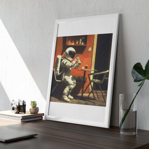 May include: A framed illustration of an astronaut sitting at a table outside, wearing a white spacesuit and holding a glass of liquid. The astronaut is sitting at a table with a red wall and a green plant behind them.