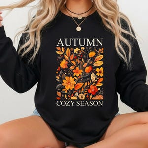 May include: A black long-sleeve sweatshirt featuring the white text AUTUMN at the top and COZY SEASON at the bottom. Centered between the text is a rectangular graphic illustration of orange, yellow, and red autumn leaves and flowers.
