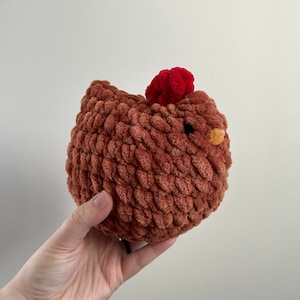May include: A brown crocheted chicken toy with a red pom-pom on its head. The chicken has a yellow beak and is made with a soft, fuzzy yarn.