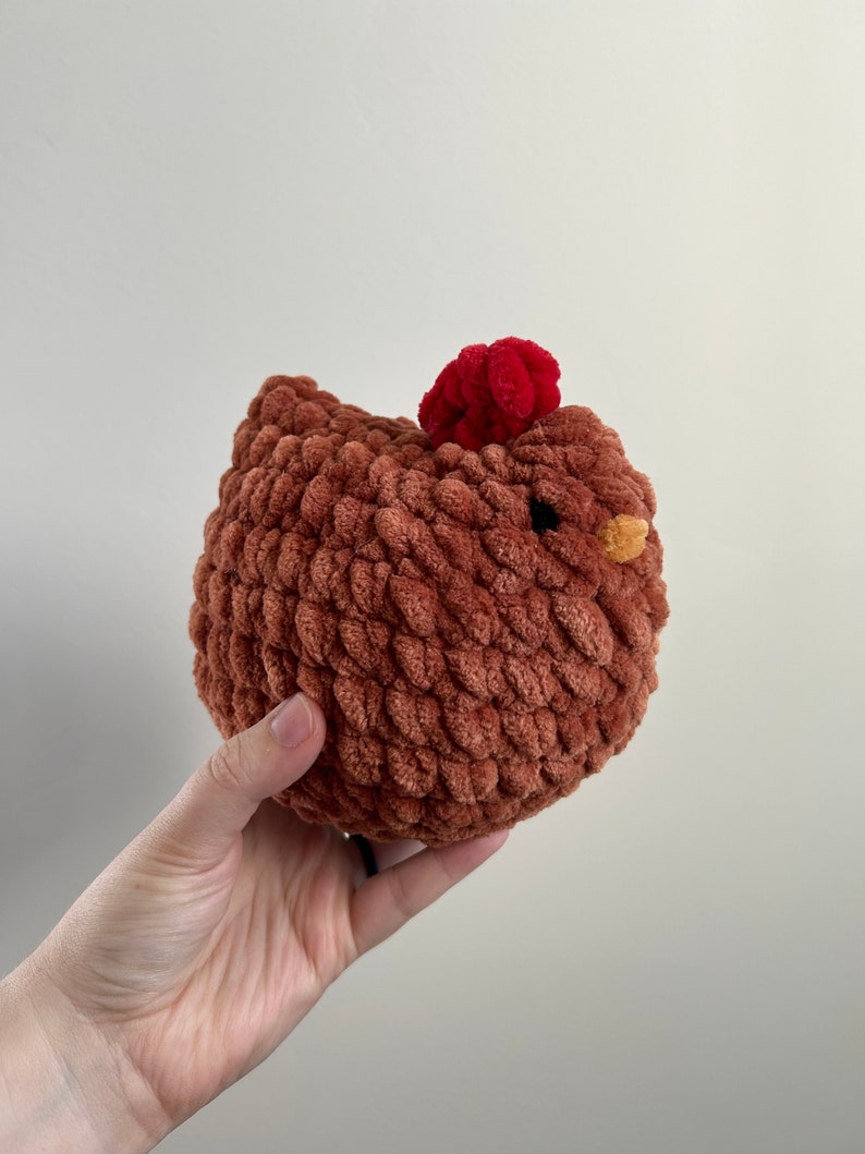 May include: A brown crocheted chicken toy with a red pom-pom on its head. The chicken has a yellow beak and is made with a soft, fuzzy yarn.