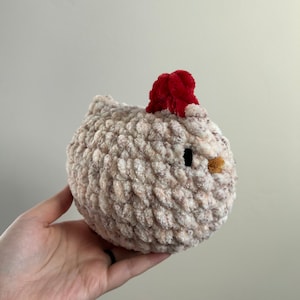 May include: A crocheted plush toy shaped like a chicken. The chicken is light brown with a red pom-pom on its head and black eyes.