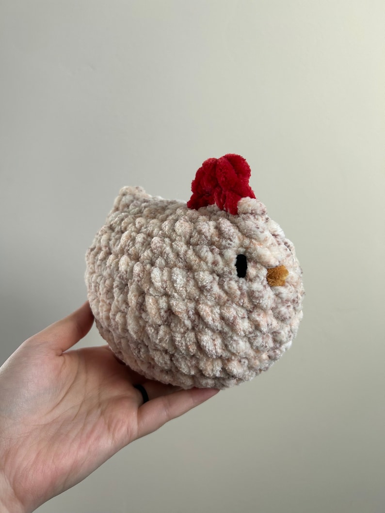 May include: A crocheted plush toy shaped like a chicken. The chicken is light brown with a red pom-pom on its head and black eyes.