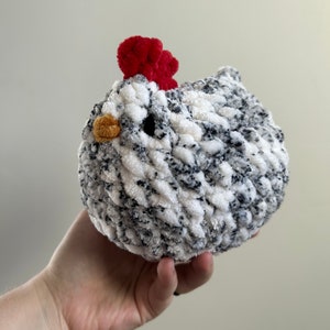 May include: A crocheted chicken toy with a white and black speckled body and a red comb. The chicken is being held in a person's hand.