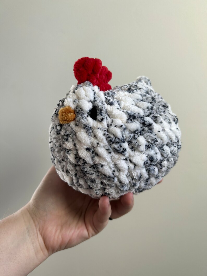 May include: A crocheted chicken toy with a white and black speckled body and a red comb. The chicken is being held in a person's hand.