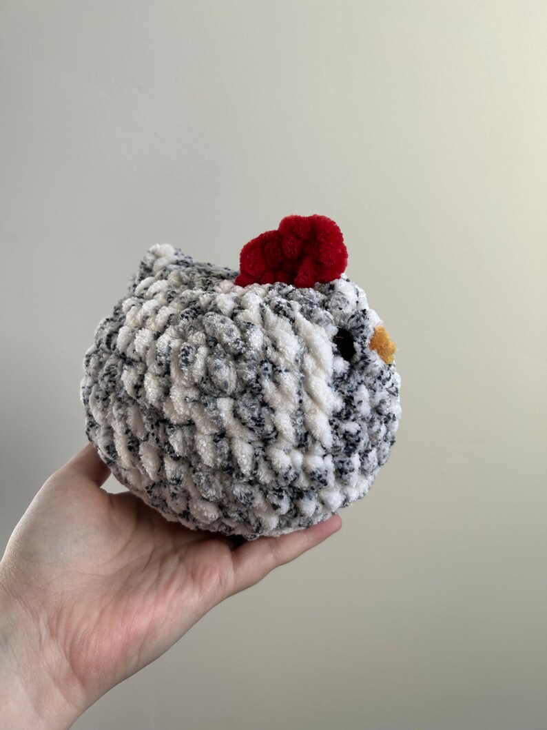 May include: A crocheted chicken toy with a white and gray speckled body, a red pom-pom for a comb, and a yellow beak.