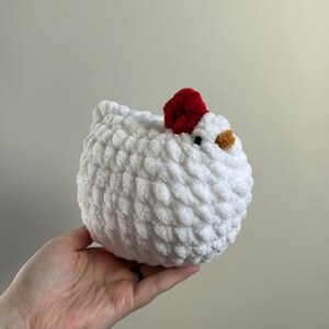 May include: A white crocheted chicken plush toy with a red pom-pom on its head. The chicken has a small, yellow beak and black eyes.