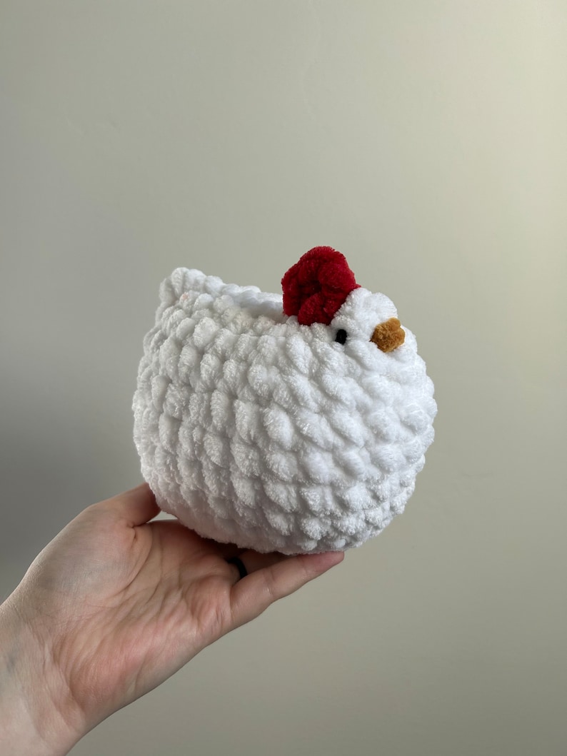 May include: A white crocheted chicken plush toy with a red pom-pom on its head. The chicken has a small, yellow beak and black eyes.