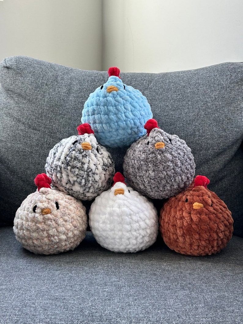 May include: Six crocheted plush chickens in different colors, including blue, gray, white, and brown, arranged in a pyramid shape on a gray couch.