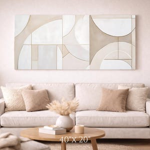 May include: Abstract wall art with a geometric design in neutral tones of beige and white. The artwork, measuring 40" x 20", hangs above a cream-colored sofa with matching pillows. A round coffee table and side table complete the living room setting.