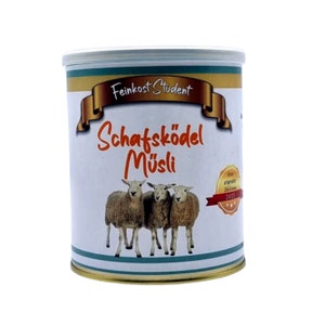Könnte beinhalten: Eine weiße Dose mit goldenem Deckel und einem blau-weißen Etikett. Das Etikett zeigt eine Karikatur von drei Schafen und den Text "Feinkost Student Schafsködel Müsli".
